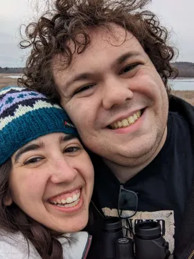 Benjamin and his partner smiling with their heads together outdoors. He has curly brown hair and wears a black sweatshirt, with binoculars at his chest. She wears a teal, cream, and lavender knit hat. A marsh and a pond are behind them under an overcast sky.